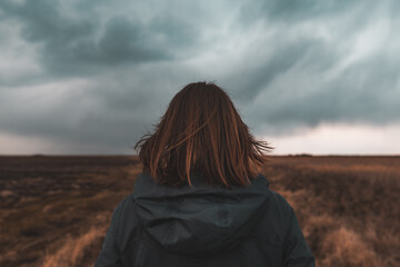 Woman standing in meadow, looking at the horizon and dark dramatic stormy clouds, rear view