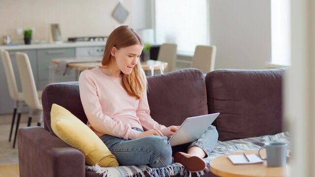 Tracking shot of beautiful young woman sitting cross-legged on sofa, working or studying on laptop from home, stretching arms and thinking