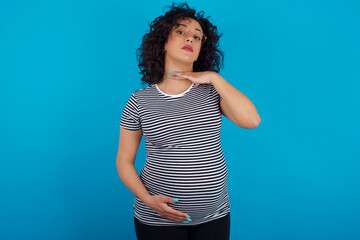 young Arab pregnant woman wearing stripped t-shirt standing against blue background cutting throat...