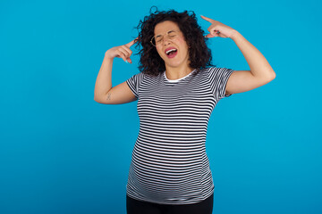 Photo of crazy young Arab pregnant woman wearing stripped t-shirt standing against blue background screaming and pointing with fingers at hair closed eyes