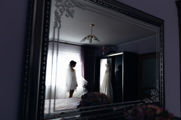 Obraz na płótnie Canvas reflections in the mirror, a slender woman in a robe with a veil stands near the bed and looks at the wedding dress.