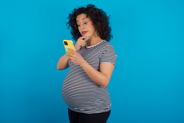 Thoughtful happy young Arab pregnant woman wearing stripped t-shirt standing against blue background hold look phone, copyspace