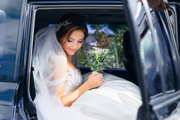 cute bride sits in a black car on the wedding day with a bouquet. Fluffy white lace dress