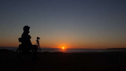 Silhouette of person in a wheelchair looking at the sunset over the ocean.  There is a camera on a tripod infront of him.