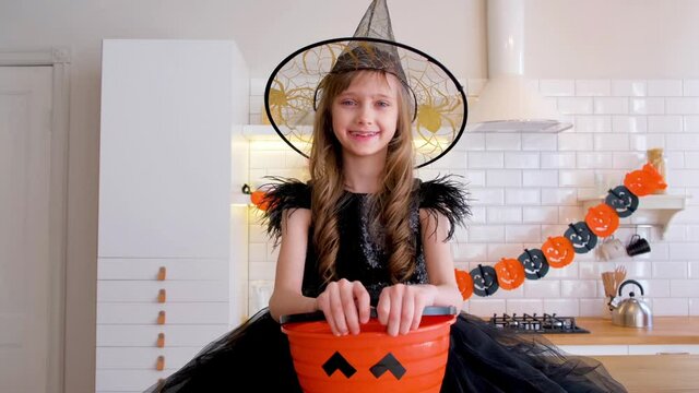 Funny blonde girl kid 8-9 years old in a witch costume sitting on the kitchen table decorated for the Halloween holiday. Traditions, holidays, scenery concept.