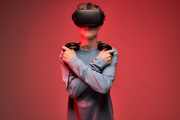 Cool teenage boy in vr goggles holding arms crossed looking forward, using controllers. isolated on red background, portrait of child engaged in video game. modern technologies concept