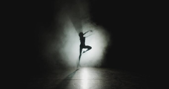 Graceful ballet female dancer jumping dancing in white smoke, floating fog in spotlight on stage. Ballerina silhouette