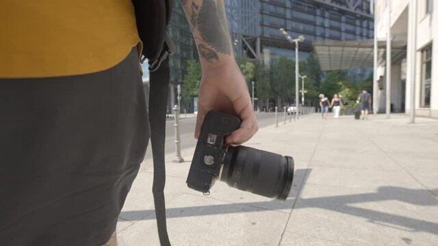 Slow Motion Close-Up Of A Man In Shorts As He Walks Along A City Sidewalk Holding A Camera On A Bright Sunny Day - Berlin, Germany
