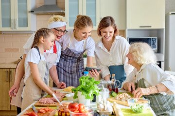 Positive beautiful women use mobile phone while cooking, watching video recipe or tips for cooking, interested in culinary. side view on family dressed in apron, at home in cozy bright kitchen