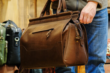 The hand of a man holding a brown travel bag against the background of other bags in the store. The concept of natural leather products.