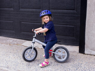 Horizontal view of cute blond toddler girl in casual clothes and helmet sitting on small bicycle in front of garage door