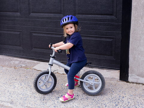 Horizontal View Of Cute Blond Toddler Girl In Casual Clothes And Helmet Sitting On Small Bicycle In Front Of Garage Door