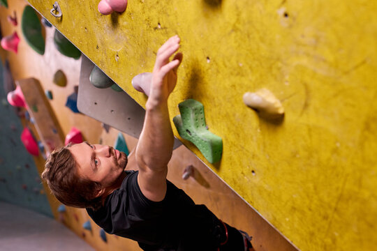 Free climber young man climbing on practical wall indoor, bouldering alone, in black sportive clothes outfit, professionally training. sport and workout concept