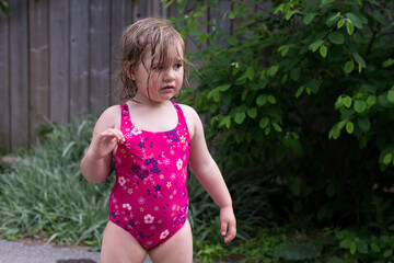 Horizontal view of cute blond dishevelled and wet chubby toddler girl wearing a pink bathing suit pouting while standing outdoors in alley