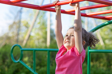 Obraz premium child and sport. little active emotional pensive girl on playground in park, hanging on horizontal bar, doing sports exercise outdoors. happy childhood. Kid play on school, kindergarten yard in summer