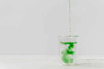 Drops of liquid chlorophyll falling down in a glass cup with water. Light white background with...