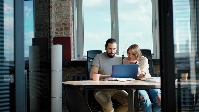 Two people coworkers a woman and a man are discussing a project while sitting in front of their laptops at the bright office.
