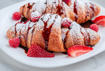 Close up of crossaints on a white plate with strawberries, raspberries and suggar glass or candy glas with chocolate syrup on top 