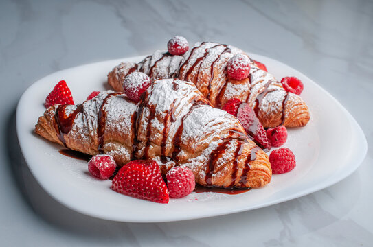 side view of croissaints or crossaints on a white plate with strawberries, raspberries, and sugar glass or candy glass with chocolate syrup on top 