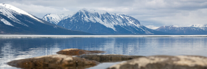 Fototapeta premium Panoramic view of a snow capped mountain landscape with reflection in water below during spring time in northern Canada in the wilderness of boreal forest. 