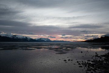 Scenic landscape in northern Canada during spring time with rocks, snow capped mountains in distance in calm lake water on cloudy pink afternoon sunset. 