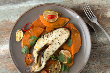 flat lay of plate of baked halibut steak with sweet potato, lime and chili on wooden table