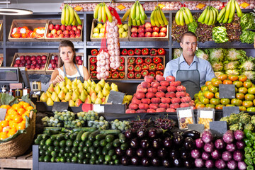 Young man and woman sellers in aprons standing near counter with the fresh vegetables and fruits on the market