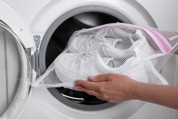 Woman putting pair of sport shoes in mesh laundry bag into washing machine, closeup