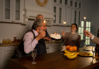Grandfather blowing out candles on birthday cake and making wish while sitting behind kitchen table at home with his wife holding cake and guests holding sparklers. Family celebration concept