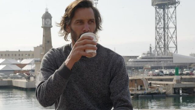 Handsome bearded man sits and sips hot drink with Barcelona Port Vell harbor in background. Spain