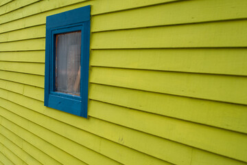 A bright yellow exterior wall of a building covered in wood clapboard siding. There's a small square glass window with blue trim and in the window a rectangular square of brown clay. 