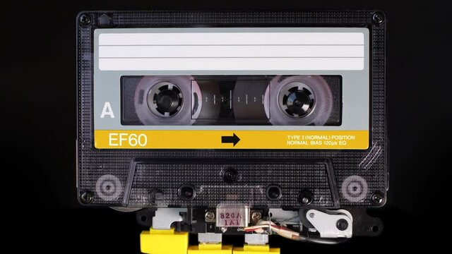 A yellow, vintage, compact cassette tape playing back in a player mechanism. Tape recorder head and rotating reels of deck player. Magnetic tape stores stereo music in a magnetic image
