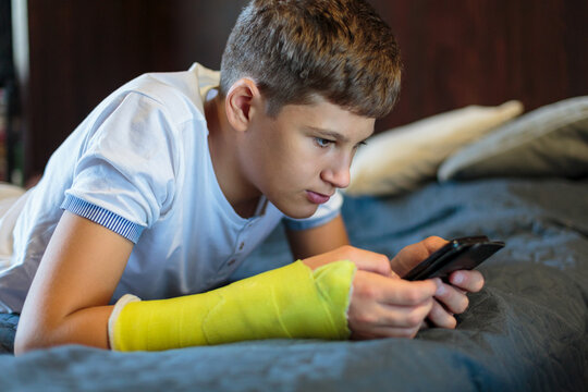 Cute boy with broken hand relaxing and smiling on couch. Close up young handsome Teenage with with yellow plaster playing smartphone at home. 