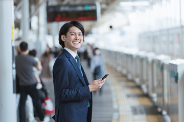 駅のホームでスマートフォンを持つ若い男性・通勤とIoTのイメージ