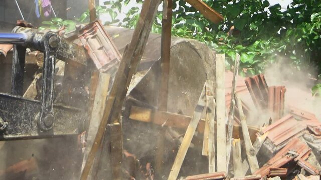 Slow motion of falling down broken roof tiles and planks during demolition of a house. Hydraulic arm pounding and crushing the roof. An excavator turns old home into rubble and dust fills the air. SM,