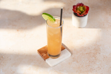 A view of a tropical yellow orange peach colored alcoholic beverage, on the rocks, with a straw and lime slice. 