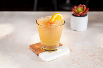 A view of a yellow orange peach colored alcoholic beverage, garnished with an orange slice and cherry, on ice. 