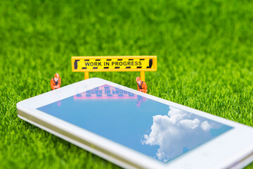 Miniature worker with smartphone and work in progress sign on green grass background, repair and...