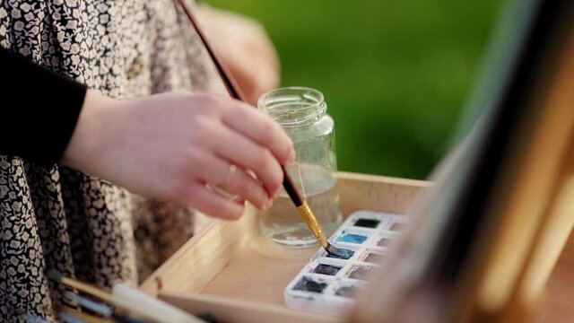 Closeup of female artist taking brush and painting with colorful watercolors in nature