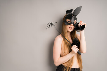 Sexy woman in a leather black fetish mask of a hare with a whip in her hands and handcuffs for sex. Stands on a gray background - obrazy, fototapety, plakaty