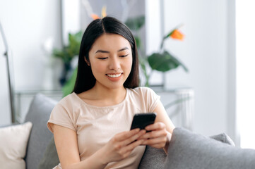 Joyful japanese attractive girl using her smartphone while sitting on sofa in stylish clothes,...