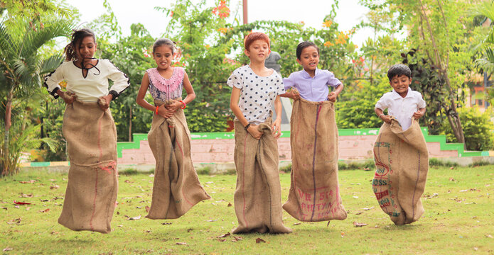 Group of Childrens playing potato sack jumping race at park outdoor during summer camp - kids having fun while playing gunny sack racing competition.