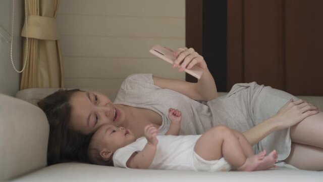 asian woman mother parent using mobile smart phone taking selfie photo with baby infant on bed at home. family video call via smartphone.  