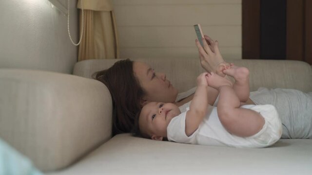asian woman mother laying down on sofa using mobile smartphone with baby infant playing beside. using mobile phone with baby at home. authentic lifestyle. life at home.
