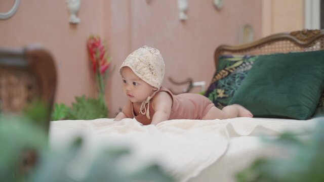 fashion portrait asian baby infant wearing hat crawling on classic vintage bed in tropical pastel pink room. baby smiling at camera and playing joyfully. kid learn to crawl improve skill development.