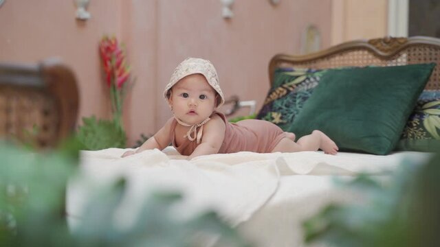 fashion portrait asian baby infant wearing hat crawling on classic vintage bed in tropical pastel pink room. baby smiling at camera and playing joyfully. kid learn to crawl improve skill development.