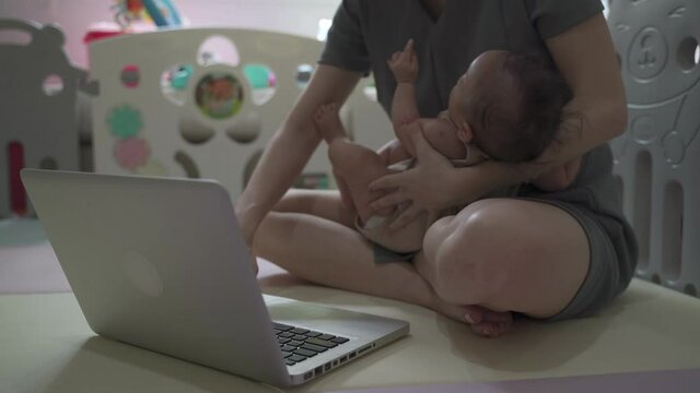 asian woman mother holding baby infant and working on computer laptop in baby playpen. working from home using computer notebook and lull baby. authentic life at home.