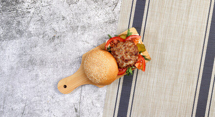 Homemade burger with pickles, herbs, onions on a small cutting wooden board on a dark grey background. Top view, flat lay