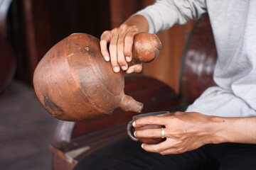 men's hands holding pots and cups made in clay. traditional Javanese kitchen tools used for warm drinks. Indonesian red kitchen utensil on a wooden background