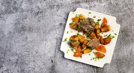 Stewed duck with baked potatoes and carrots on a square plate on a dark grey background. Top view, flat lay